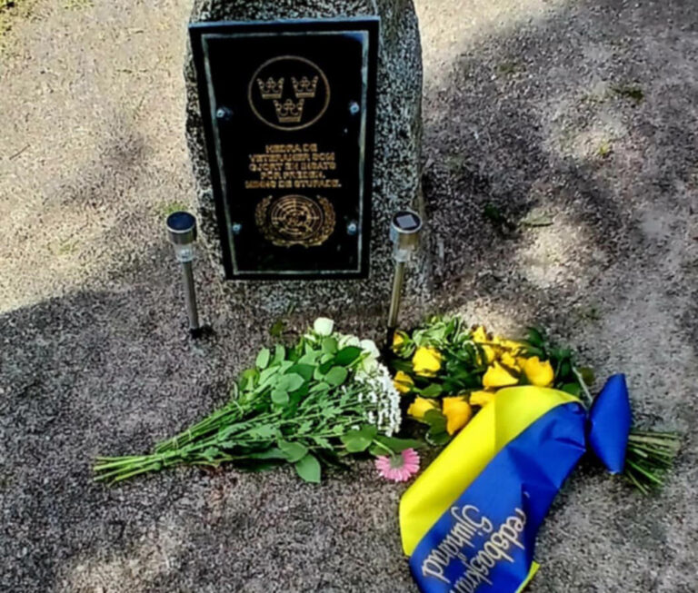 Två blombuketter ligger på marken framför en sten med minnesplakett.